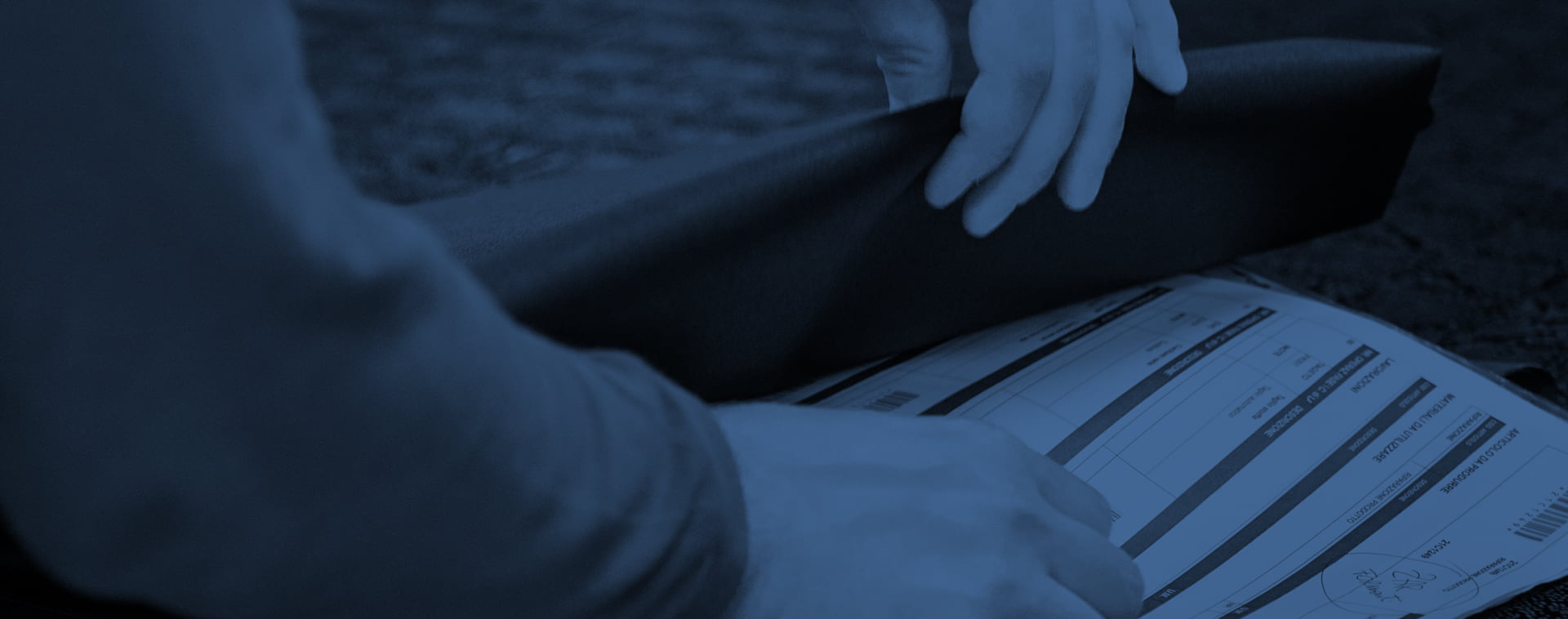Primo piano di mani che tengono un tessuto nero arrotolato e diversi fogli di carta o documenti stesi su una superficie. L'immagine è colorata di blu scuro.